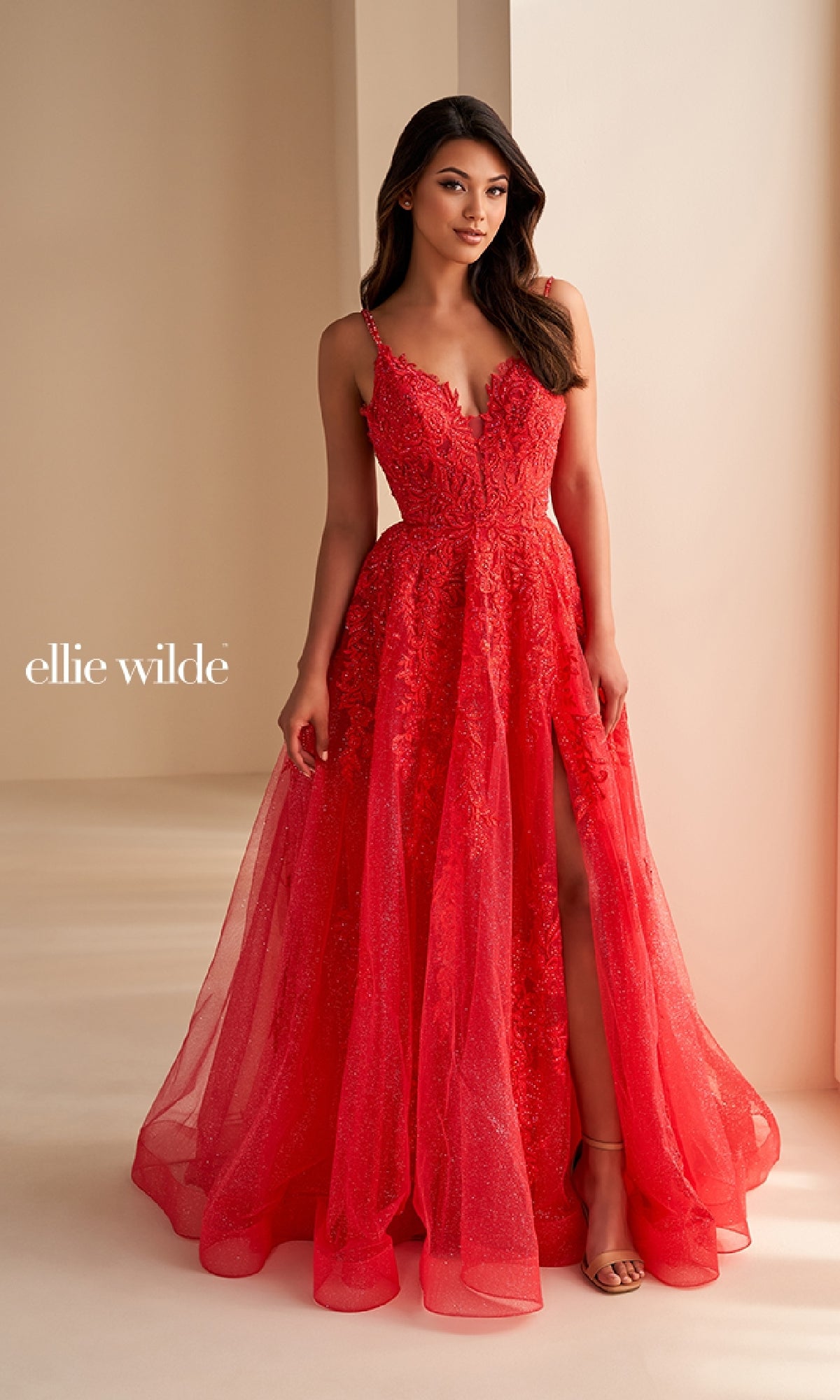 A woman indoors models the Ellie Wilde EW36226 long prom dress in bright red, featuring an A-line silhouette, lace details, corset bodice, and thigh-high slit. The